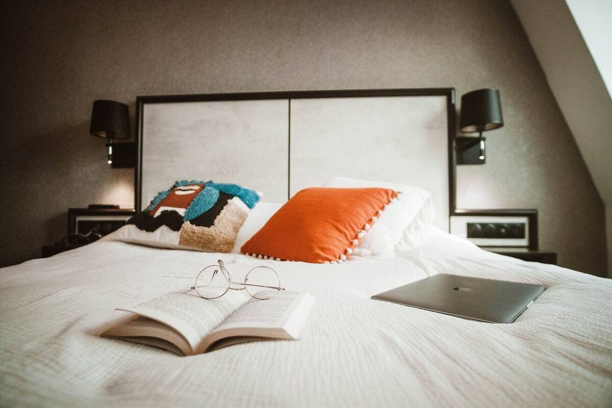 A neatly made bed with a laptop, glasses, and an open book, a colorful blanket, and two pillows.
