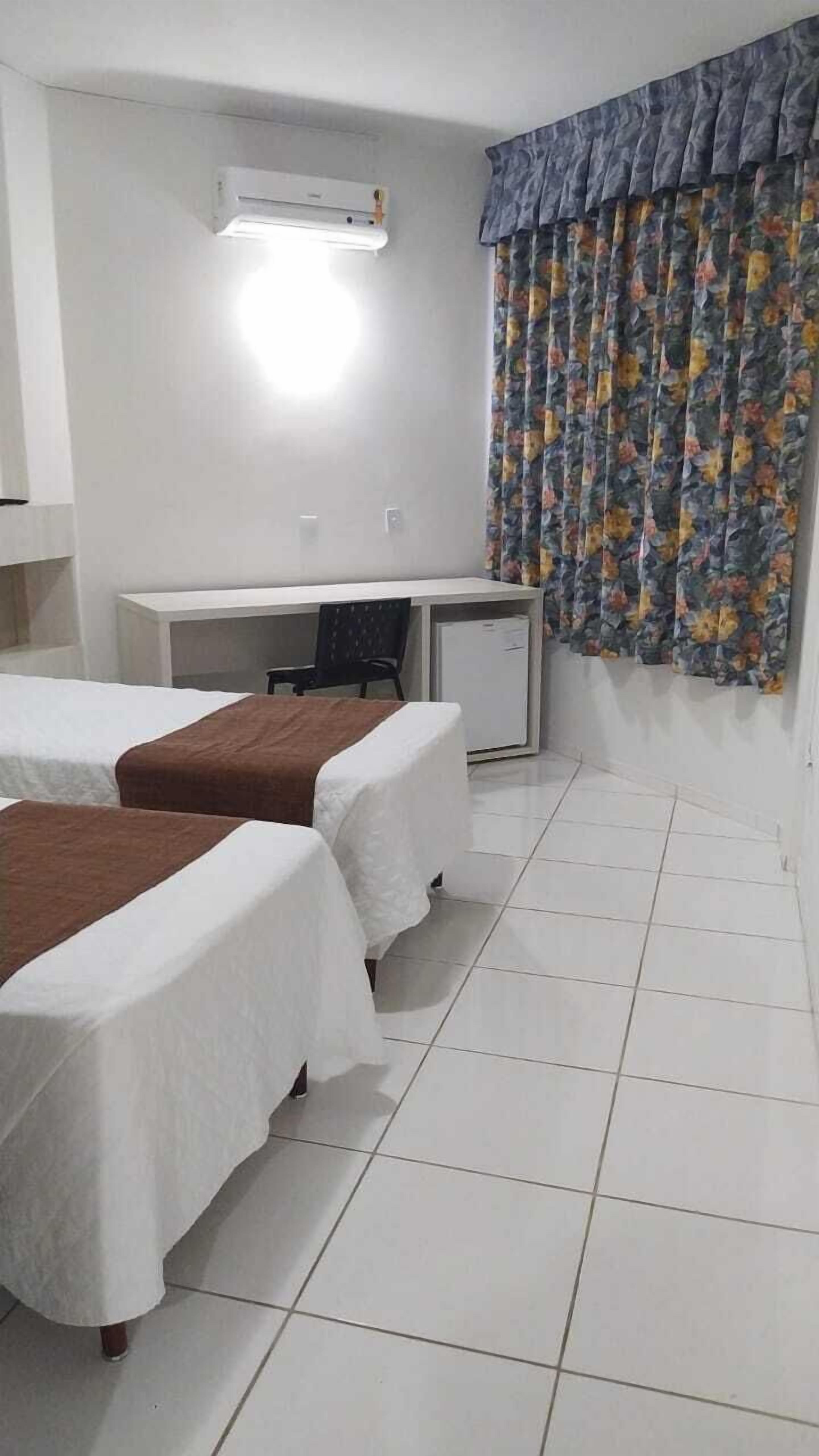 A hotel room with two beds, a desk, a chair, and a window with floral curtains.