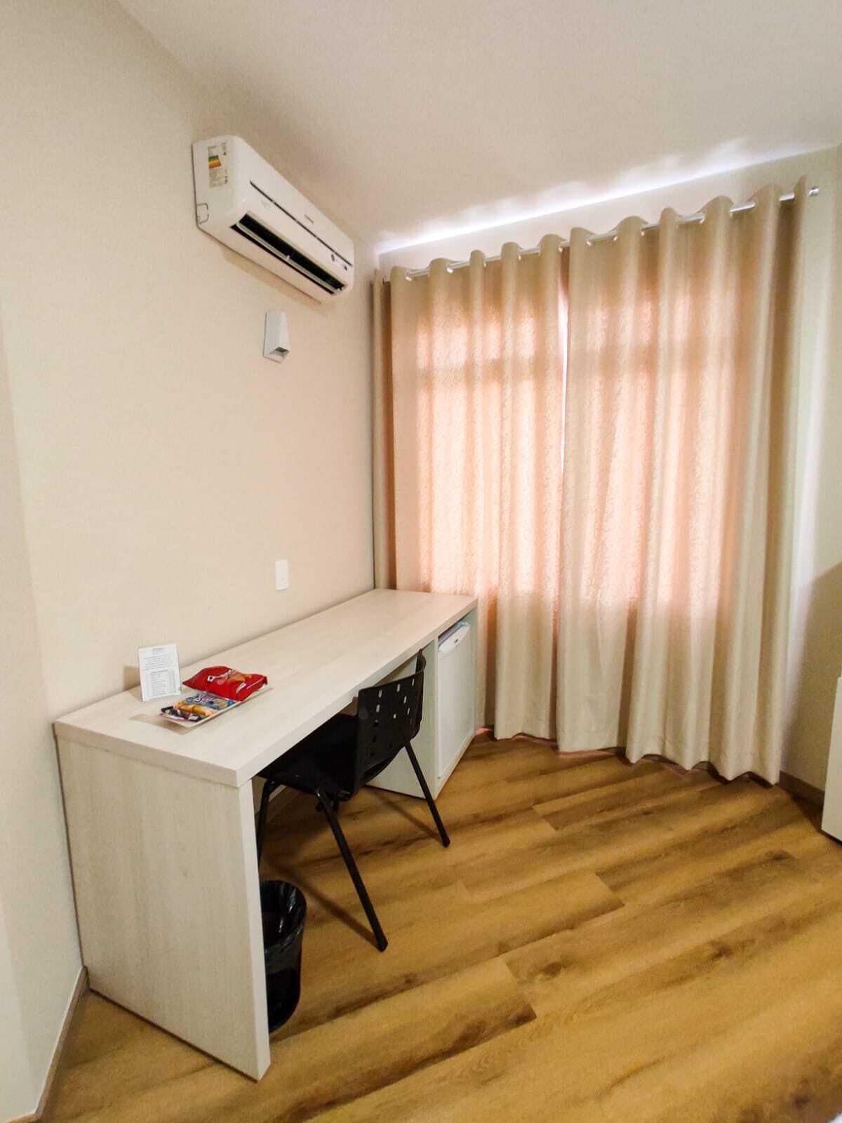 A small, neatly arranged room with a desk, a chair, a trash bin, and a window with curtains.