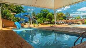 A swimming pool with a blue slide, lounge chairs, and a covered area with a view of trees and a clear sky.