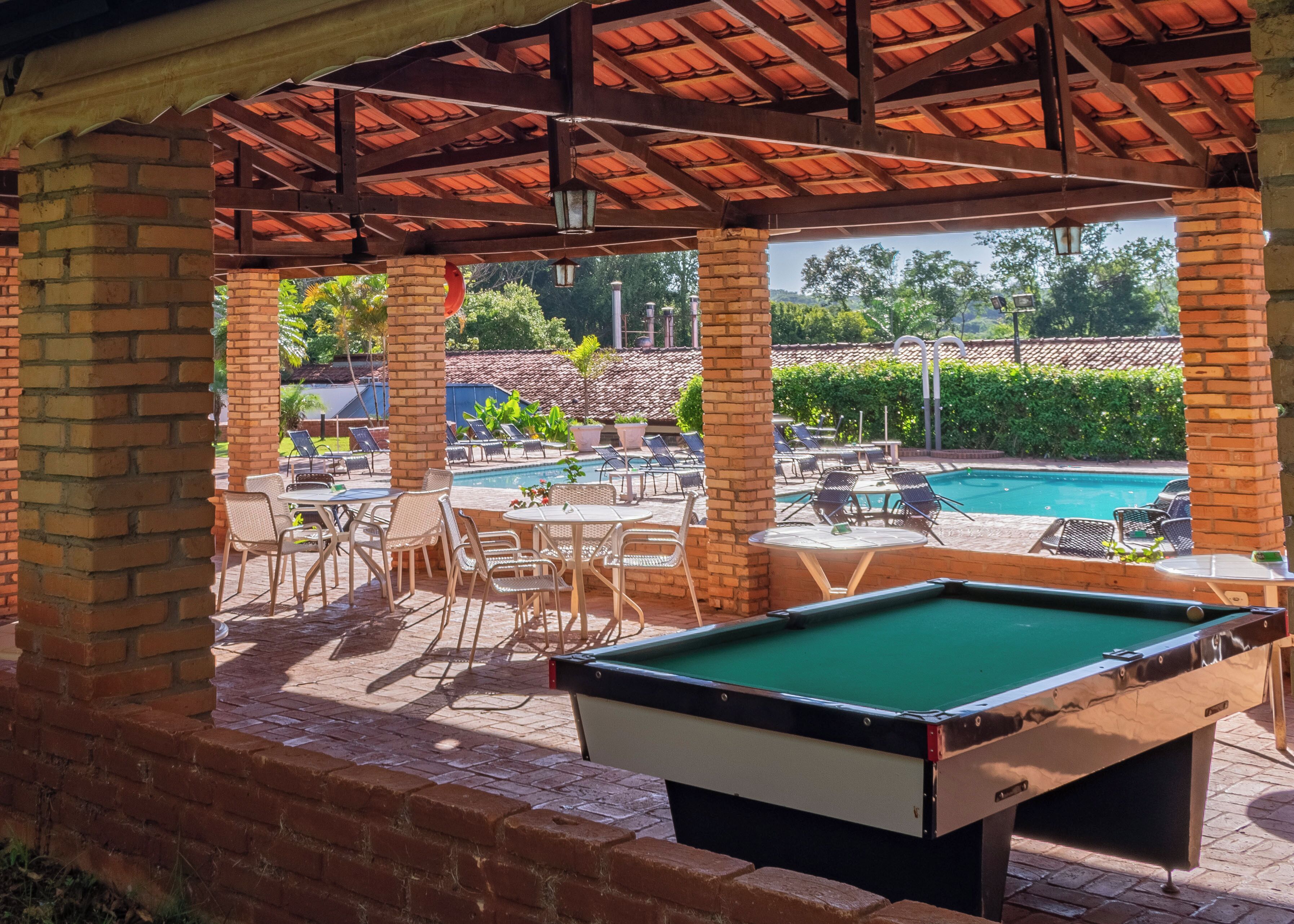 Un área de piscina con mesa de billar, sillas de descanso y una zona de estar cubierta con techo de baldosas.
