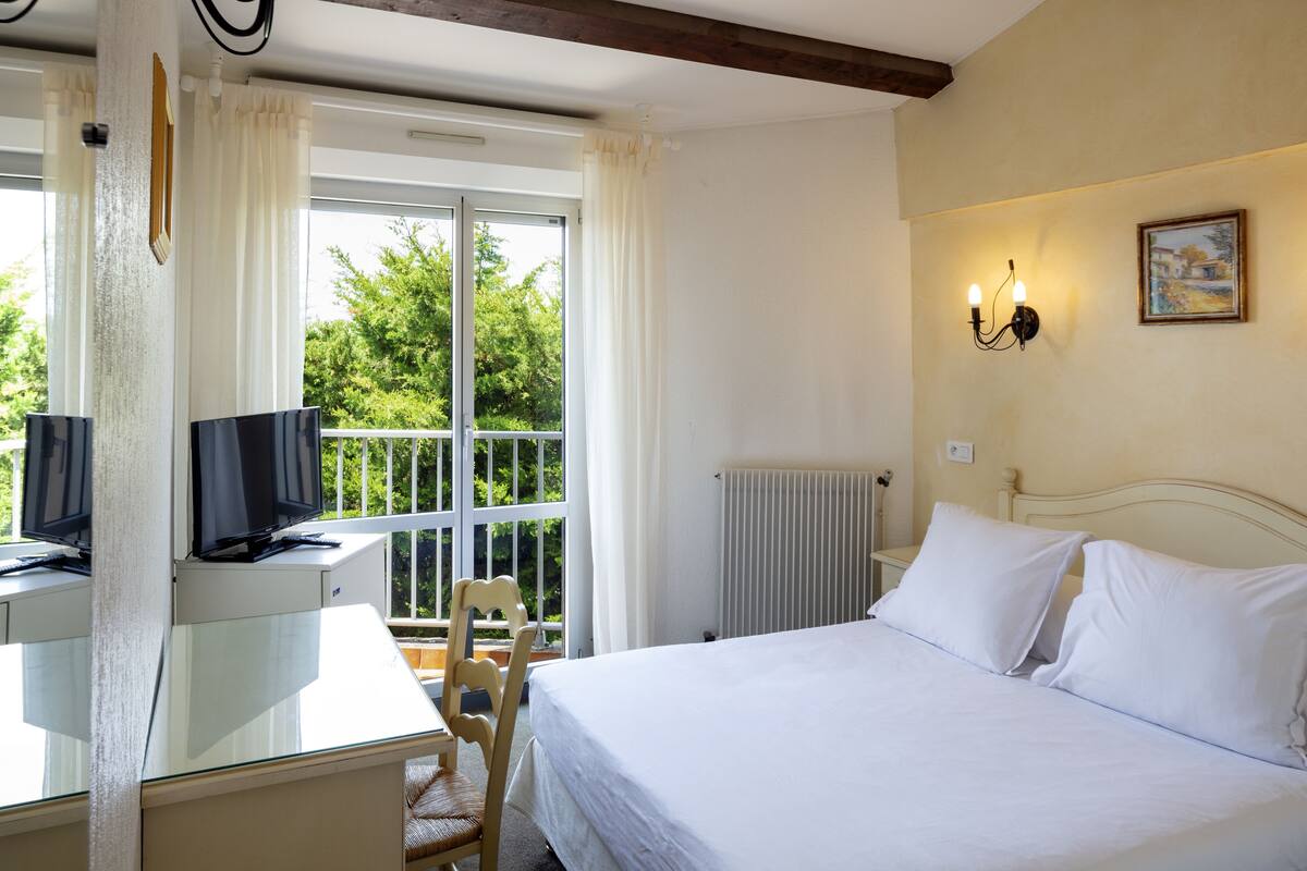 A hotel room with a bed, a desk with a television, a chair, a window with curtains, and a wall-mounted light fixture.
