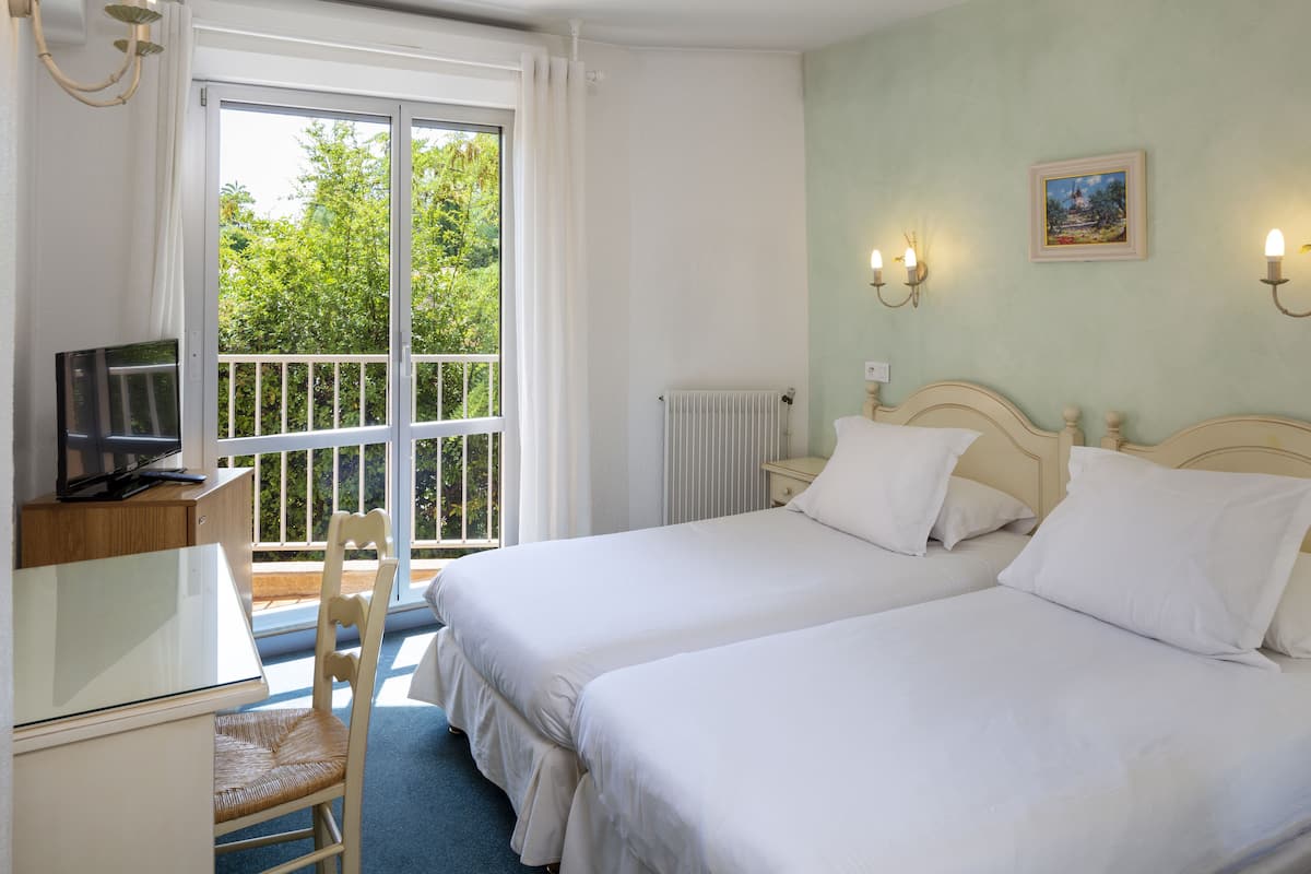 A hotel room with two beds, a desk with a television, a chair, and a view of greenery through a large window.