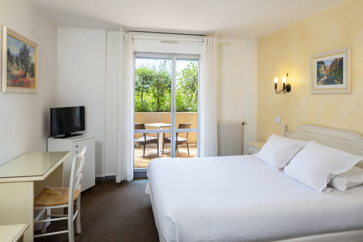 A hotel room with a bed, a desk with a television, a chair, and a view of the outdoors through a sliding door.
