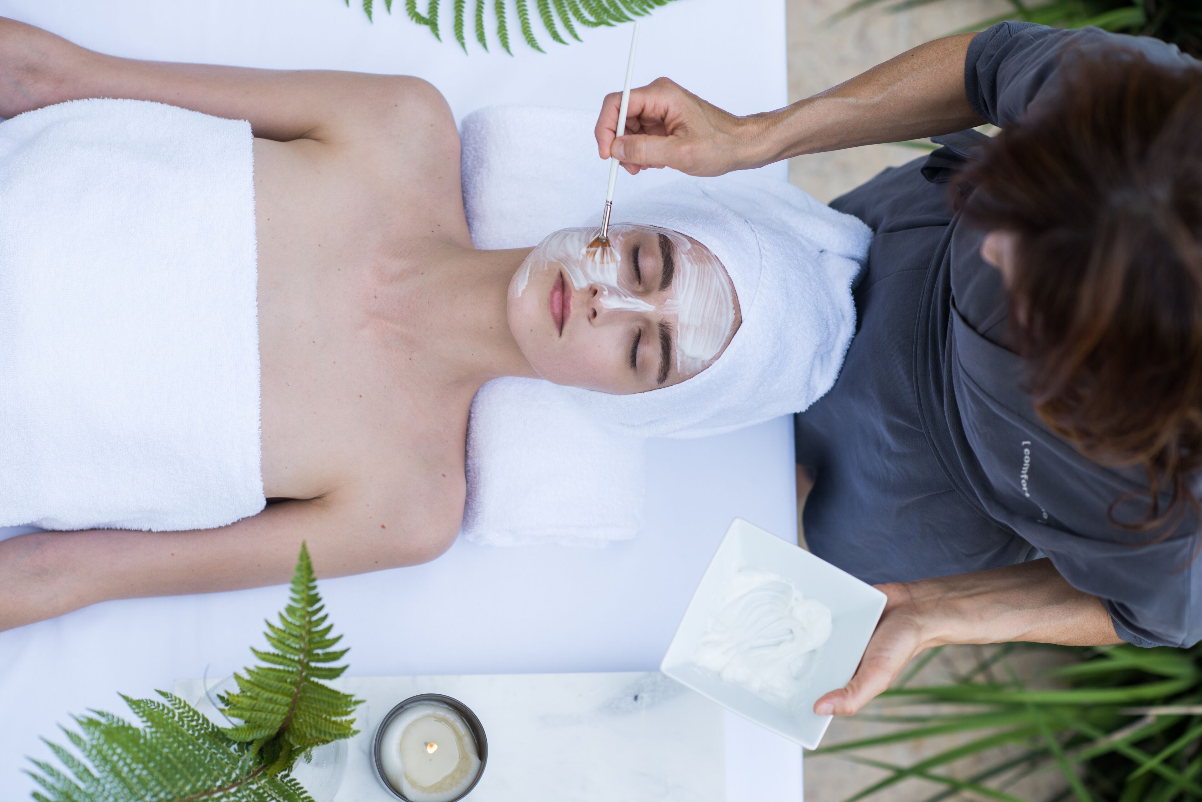 Une personne reçoit un soin du visage, une serviette est enroulée autour de sa tête, une bougie et une plante sont posées sur une table.