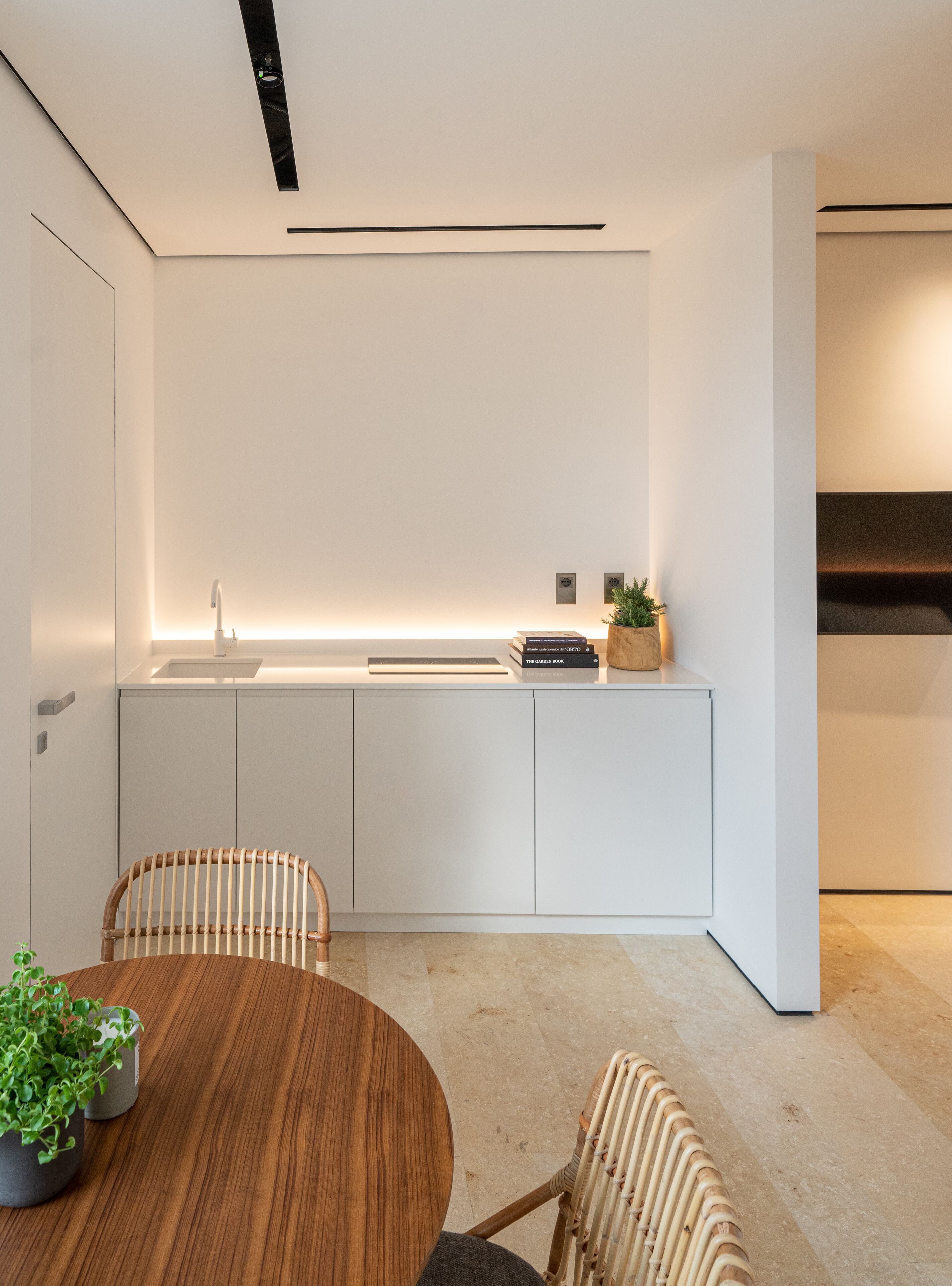 A modern kitchen with white cabinetry, a built-in oven, and a countertop with a sink and books. There is a round wooden table with two wicker chairs and a potted plant.