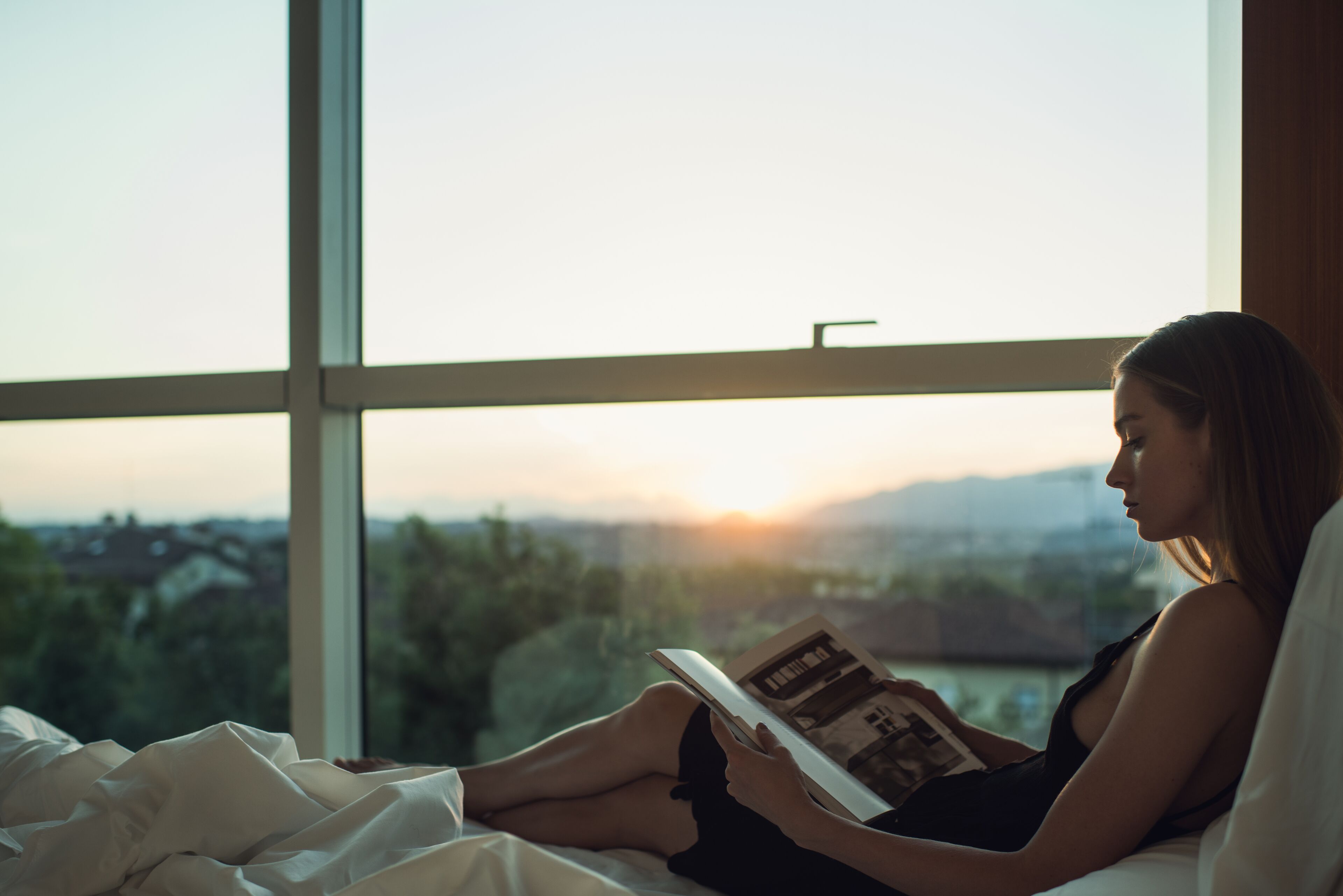 A person sitting on a bed reading a book by a window with a view of a cityscape and mountains.