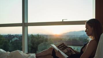 A person sitting on a bed reading a book by a window with a view of a cityscape and mountains.