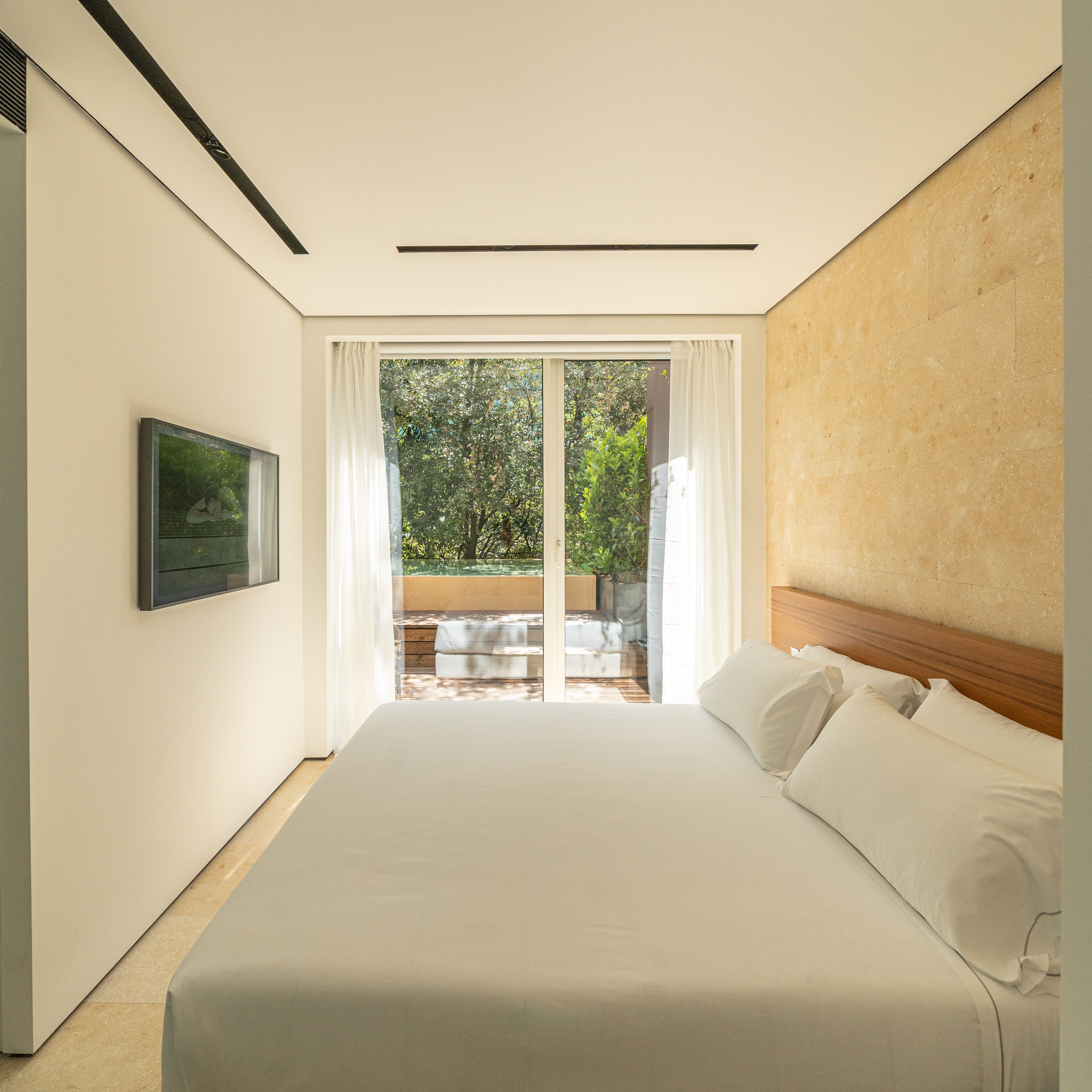 A modern bedroom with a large bed, a TV mounted on the wall, and a sliding glass door leading to an outdoor area.