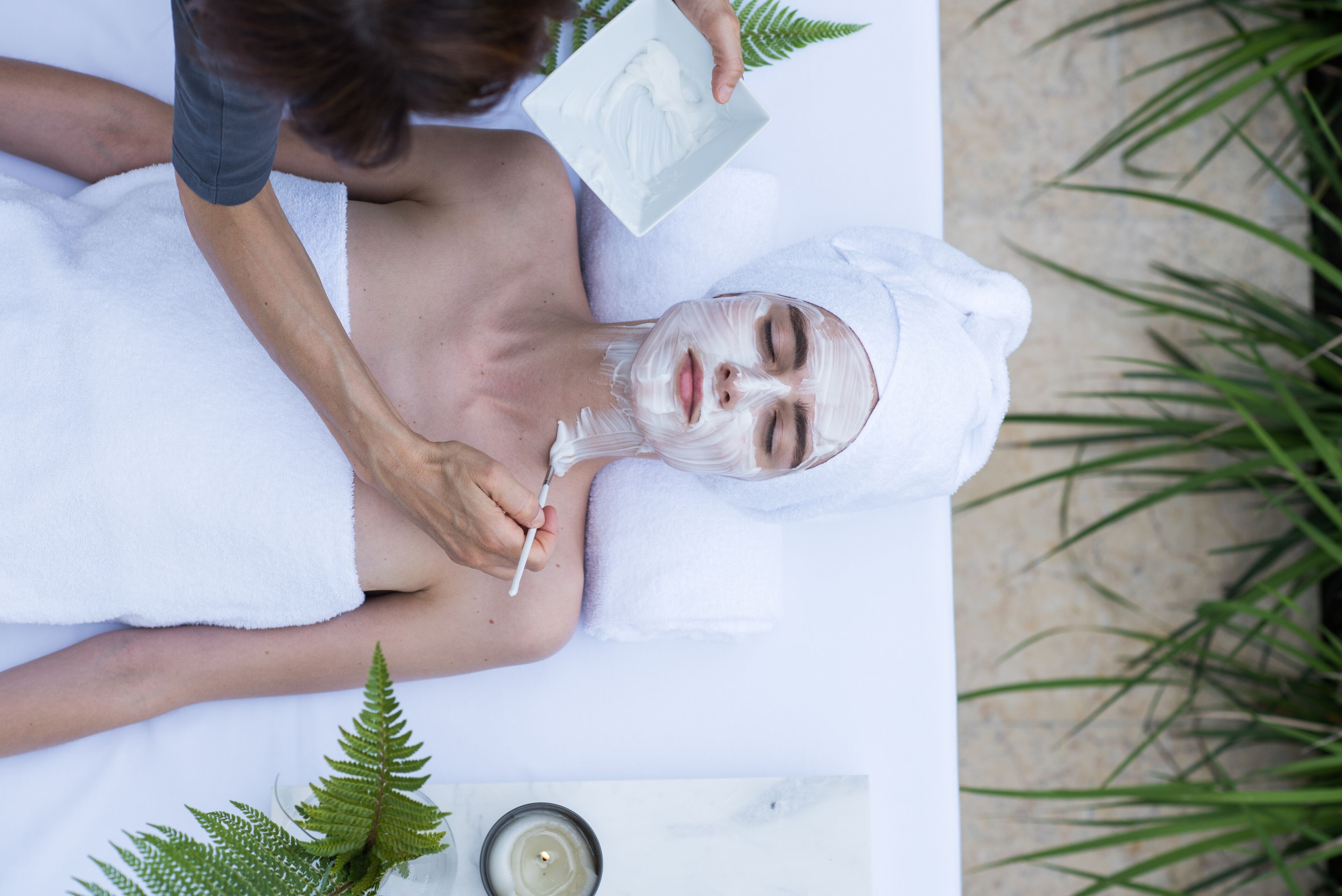 A person receiving a facial treatment in a spa setting with a white towel, a plant, and a candle on a table.