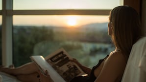 Una persona leyendo un libro junto a una ventana con vistas a la puesta de sol.
