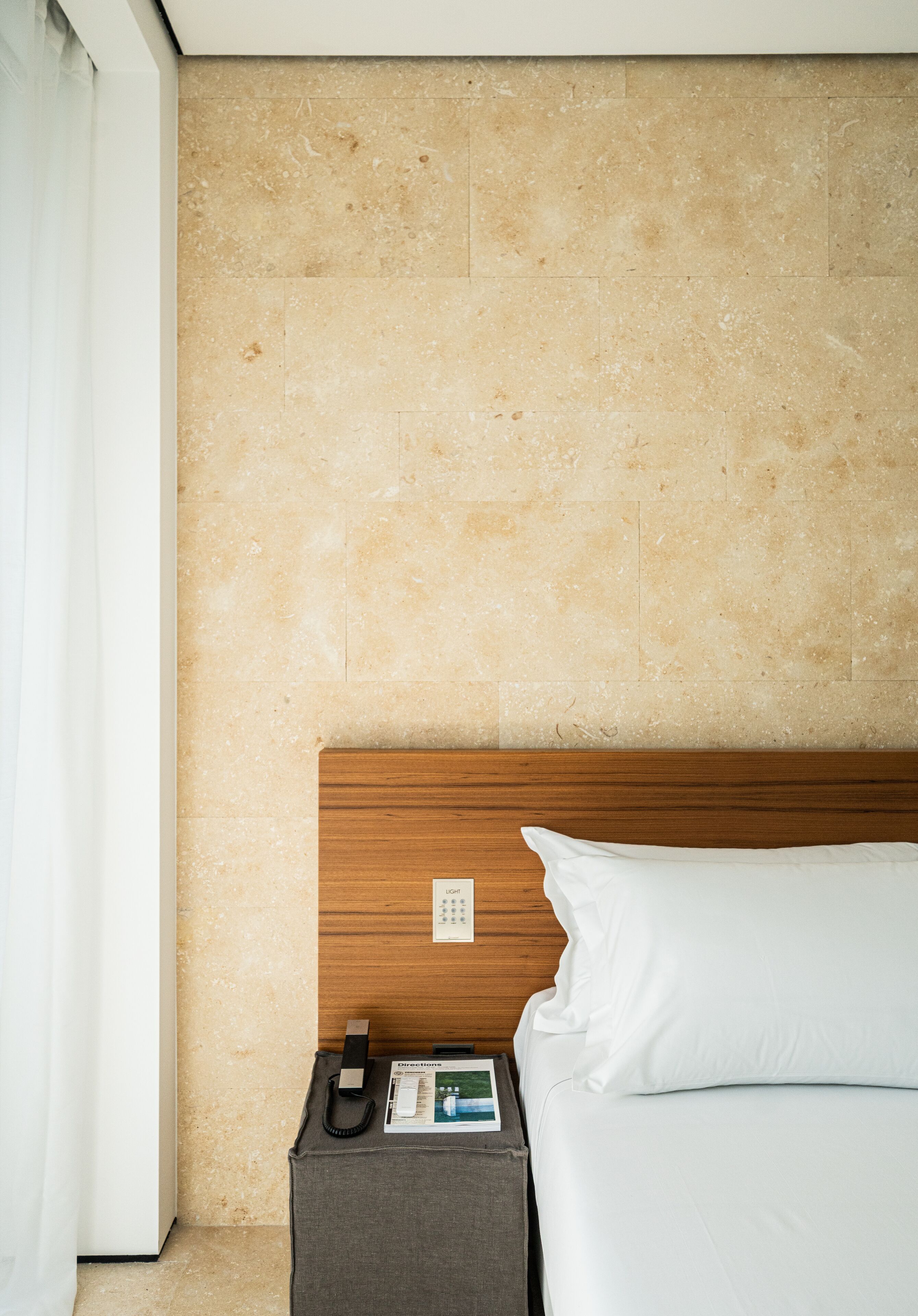 Una camera d'hotel con una testiera in legno, un letto con biancheria da letto bianca, un comodino con un telefono e una brochure, e un muro rivestito di piastrelle.