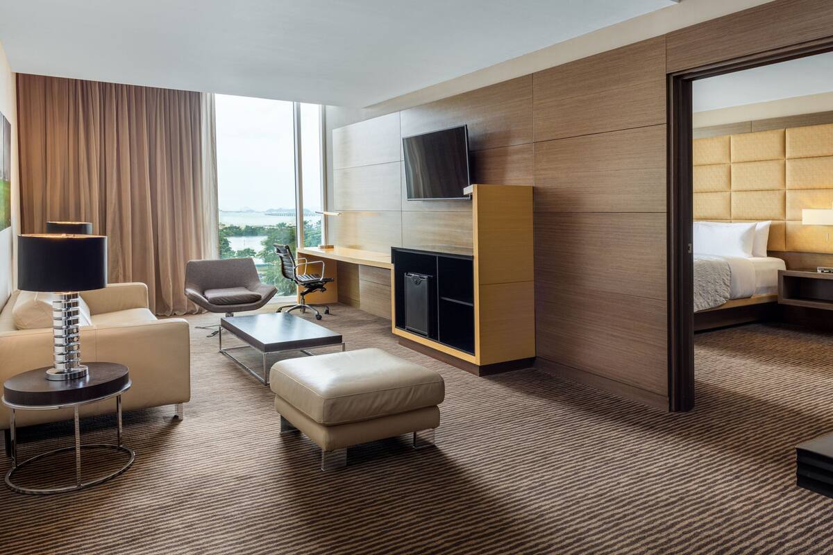 A modern hotel room with a sofa, armchair, ottoman, flat-screen TV, fireplace, and a bed with a headboard.