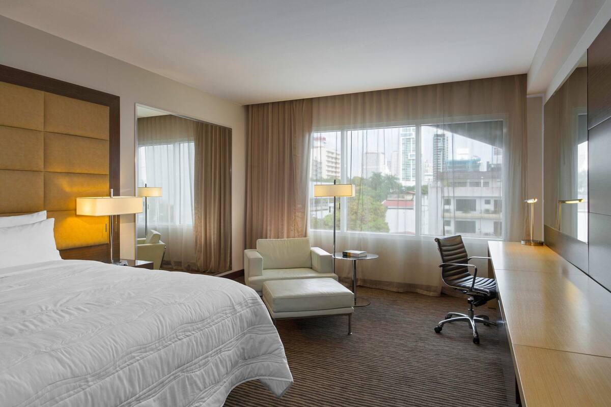 A hotel room with a large bed, a desk, a chair, and a view of the city through the window.