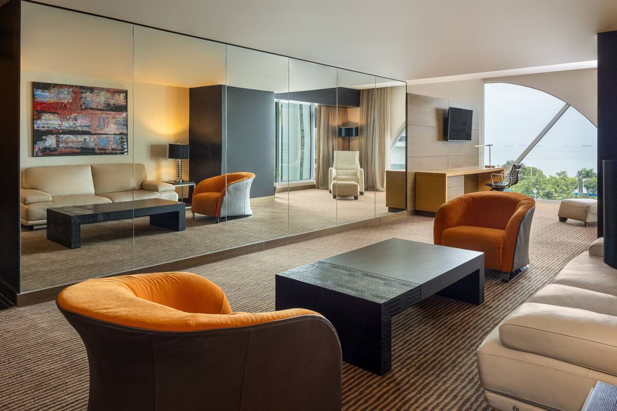 A modern hotel room with a sofa, armchairs, a coffee table, a TV, and a large window offering a view.