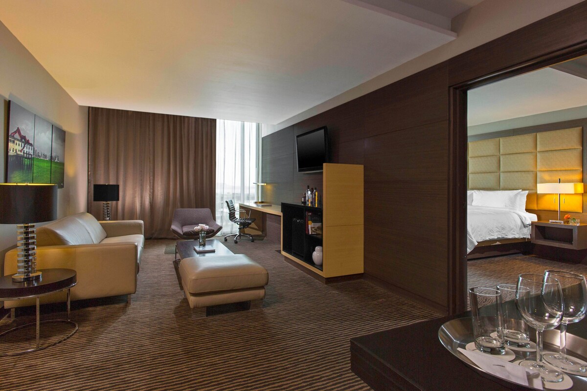 A hotel room with a sofa, armchair, ottoman, flat-screen TV, bedside table, and a bed with a headboard.