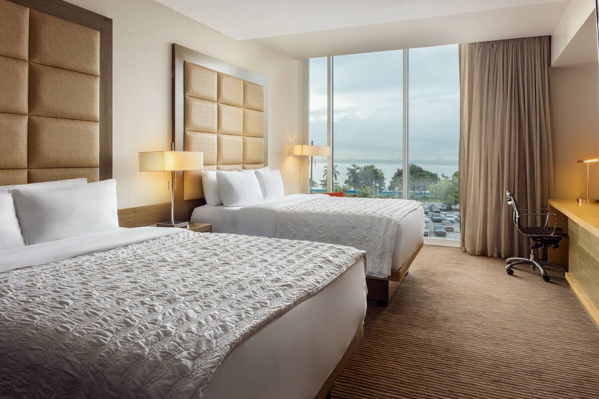 A hotel room with two beds, a large window with a view, a desk, and a chair.