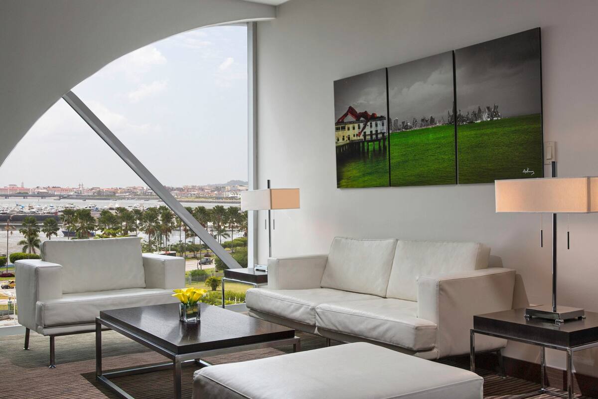 A modern hotel room with a large window offering a city view, a white sofa, a coffee table, and a painting on the wall.