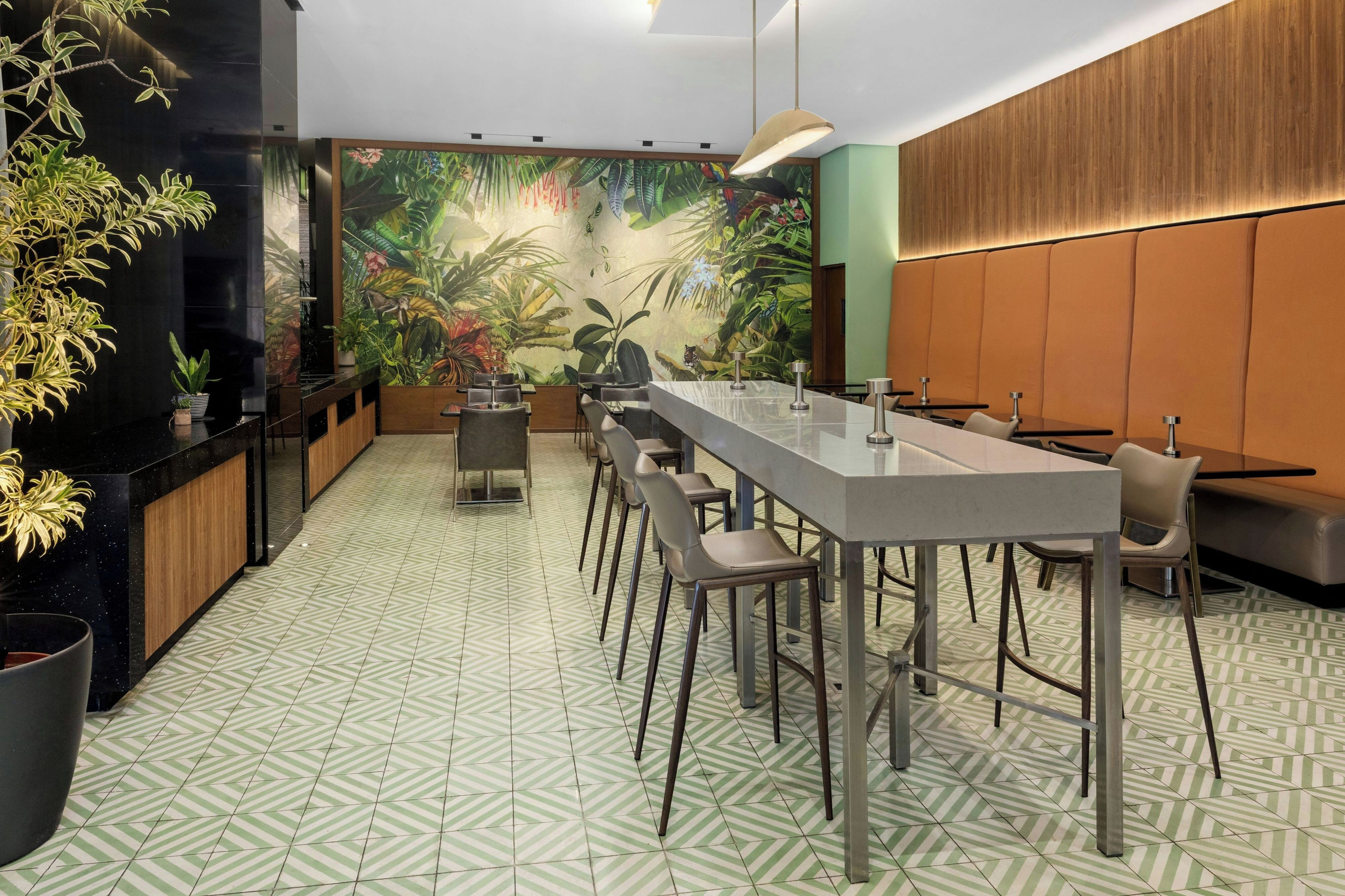 Un bar moderno con una barra larga, diversas opciones de asientos y un mural tropical en la pared.