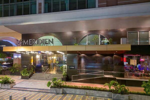 A hotel entrance with a modern design, featuring a prominent 'Le Meridien' sign, a glass facade, and a well-lit outdoor area with plants.