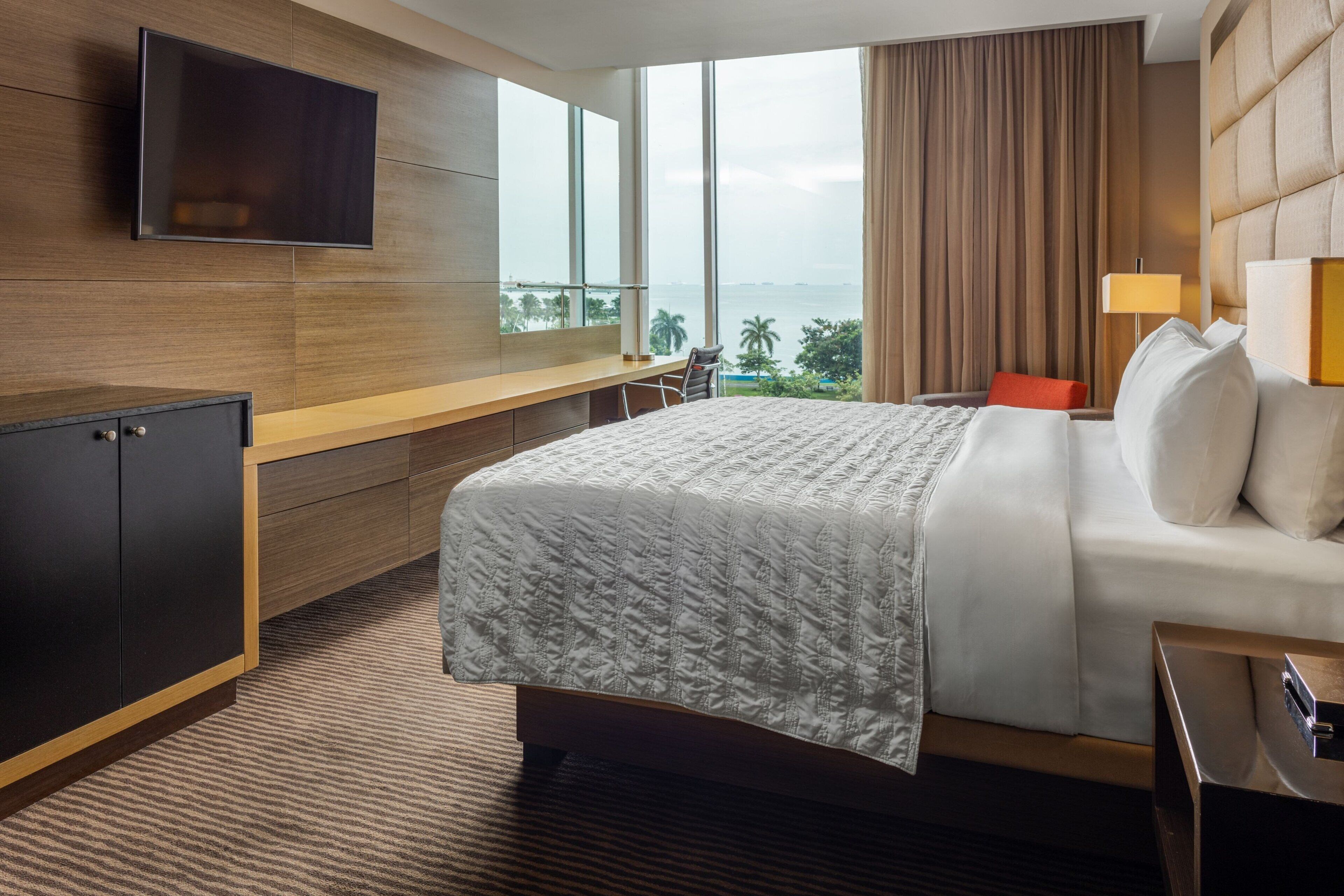Un cuarto de hotel con una cama grande, un escritorio con silla, un televisor montado en la pared y vista al mar a través de amplios ventanales.