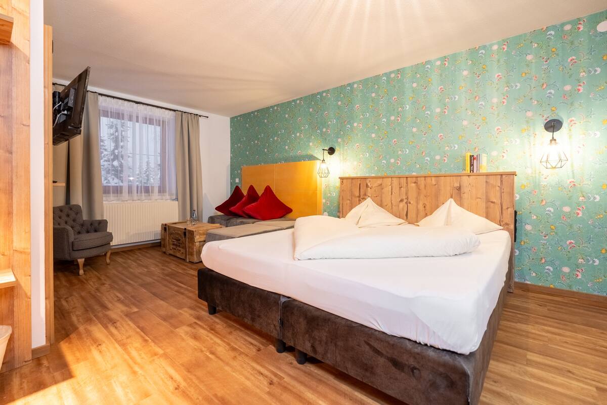 A bedroom with a wooden bed, a grey sofa, a wooden nightstand, a TV mounted on the wall, and a window with curtains.