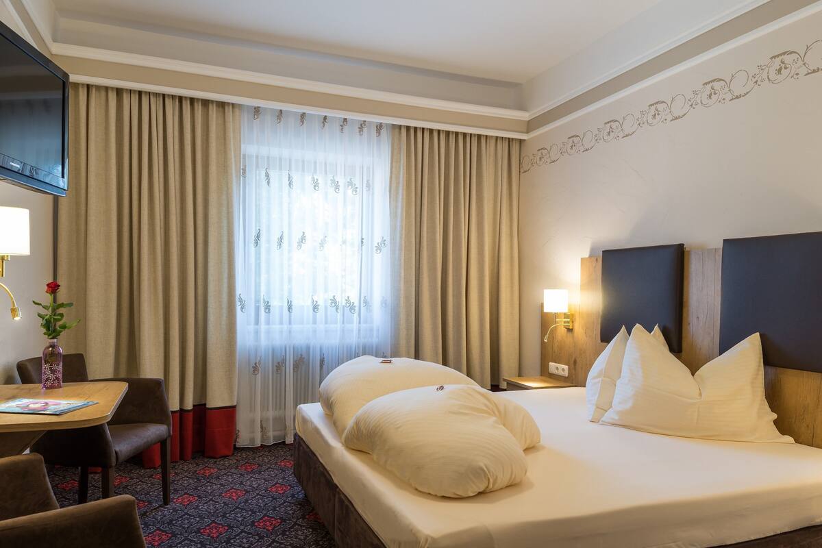 A hotel room with a large bed, a desk with a chair, a TV mounted on the wall, and a window with curtains.