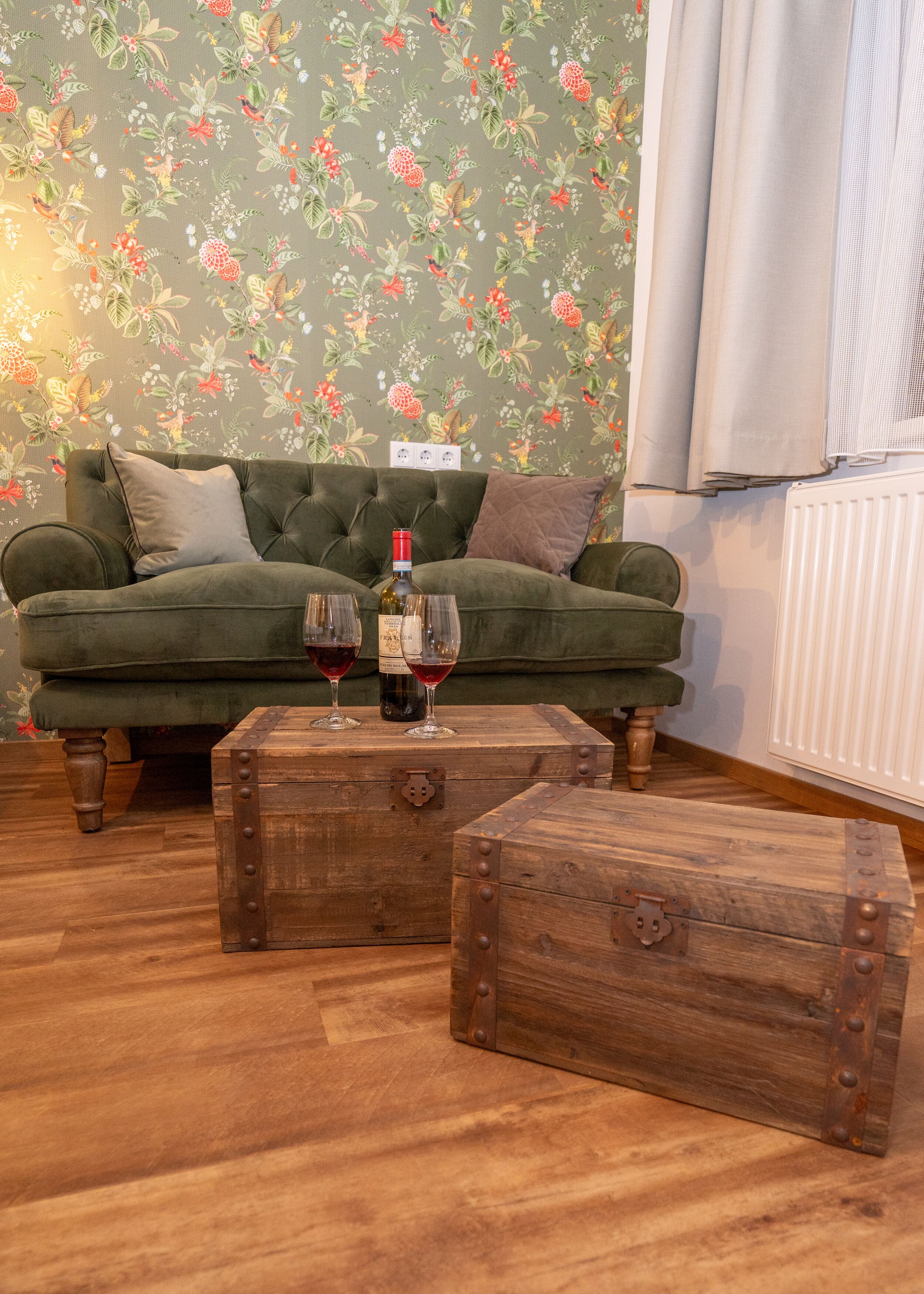 Ein grünes Sofa mit zwei Kissen, ein Couchtisch aus Holz mit einer Flasche und Gläsern sowie eine Wand mit Blumenmuster.