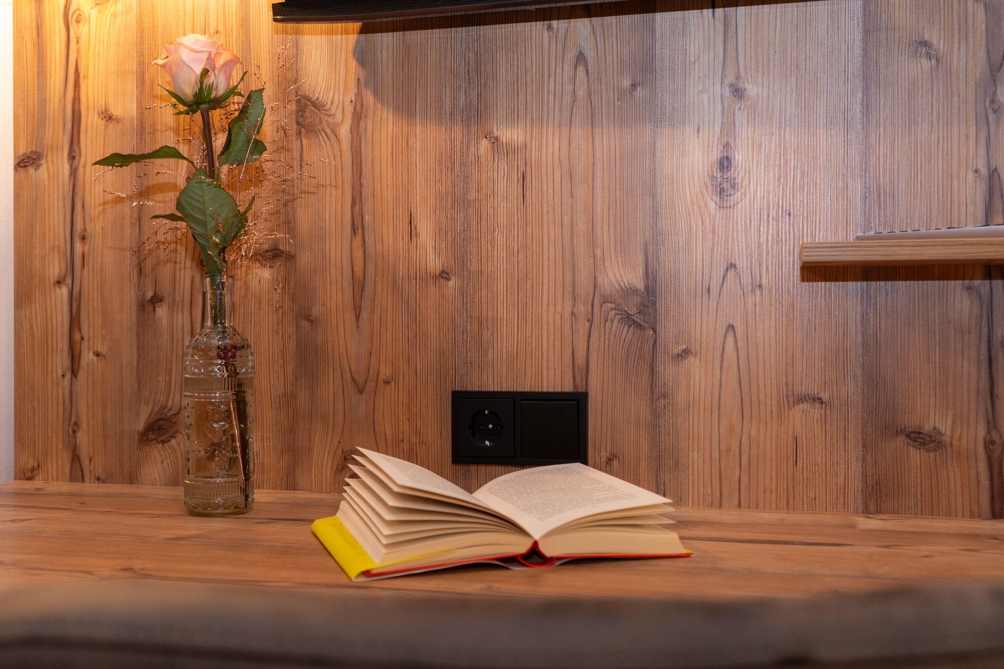 A wooden wall with a wooden shelf, a book, and a vase with a flower.