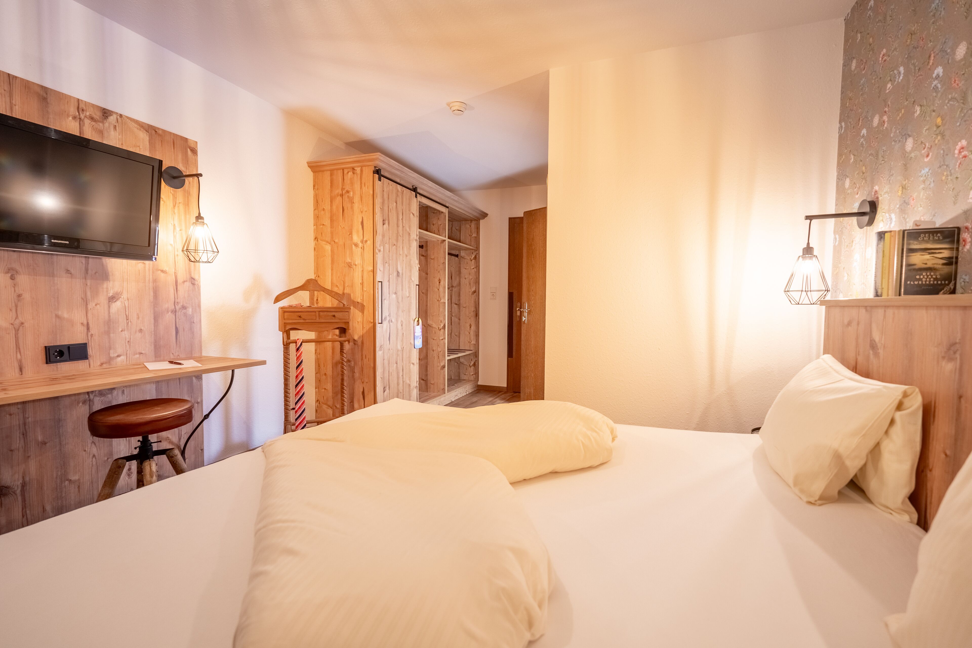 Un dormitorio con una cama, un escritorio con silla, un televisor montado en la pared y un armario.
