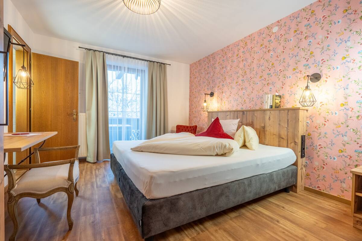 A bedroom with a large bed, wooden floor, a desk, and a window with curtains.