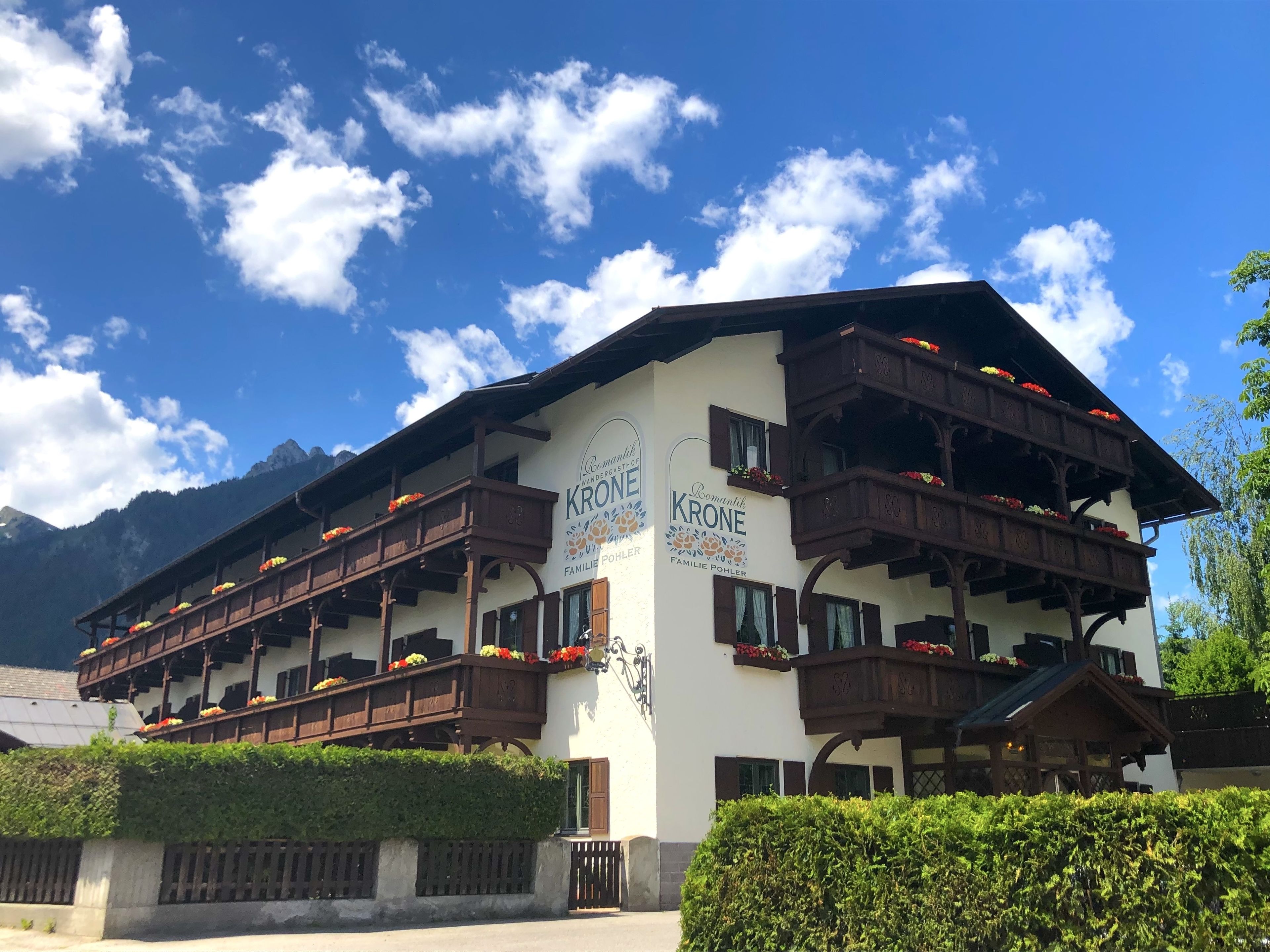 Un edificio alpino tradicional con varios balcones decorados con flores, un letrero que indica que es un negocio familiar, y un paisaje montañoso de fondo.