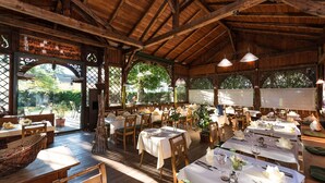Un restaurante al aire libre con mesas y sillas de madera, cubierto por un techo de madera, y grandes ventanales que ofrecen vistas de la vegetación circundante.
