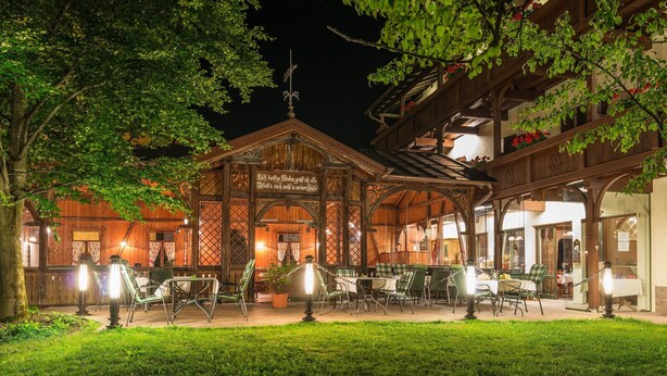 Un espacio de comedor al aire libre con mesas y sillas de madera, rodeado por un jardín y un edificio con un letrero.
