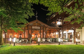 An outdoor dining area with wooden tables and chairs, surrounded by a garden and a building with a sign. - Hotel Krone Tirol (Lechaschau)