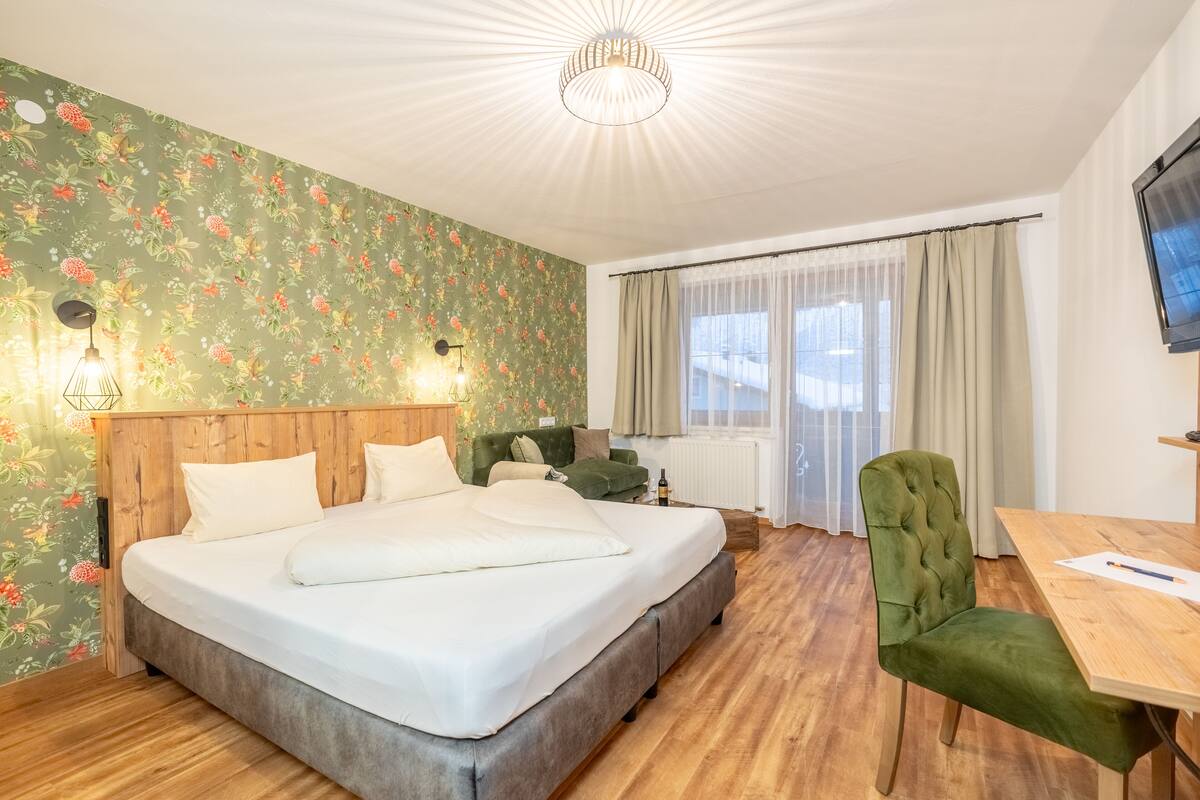 A hotel room with a large bed, a wooden headboard, a green upholstered chair, a wooden desk, and a television on the wall.