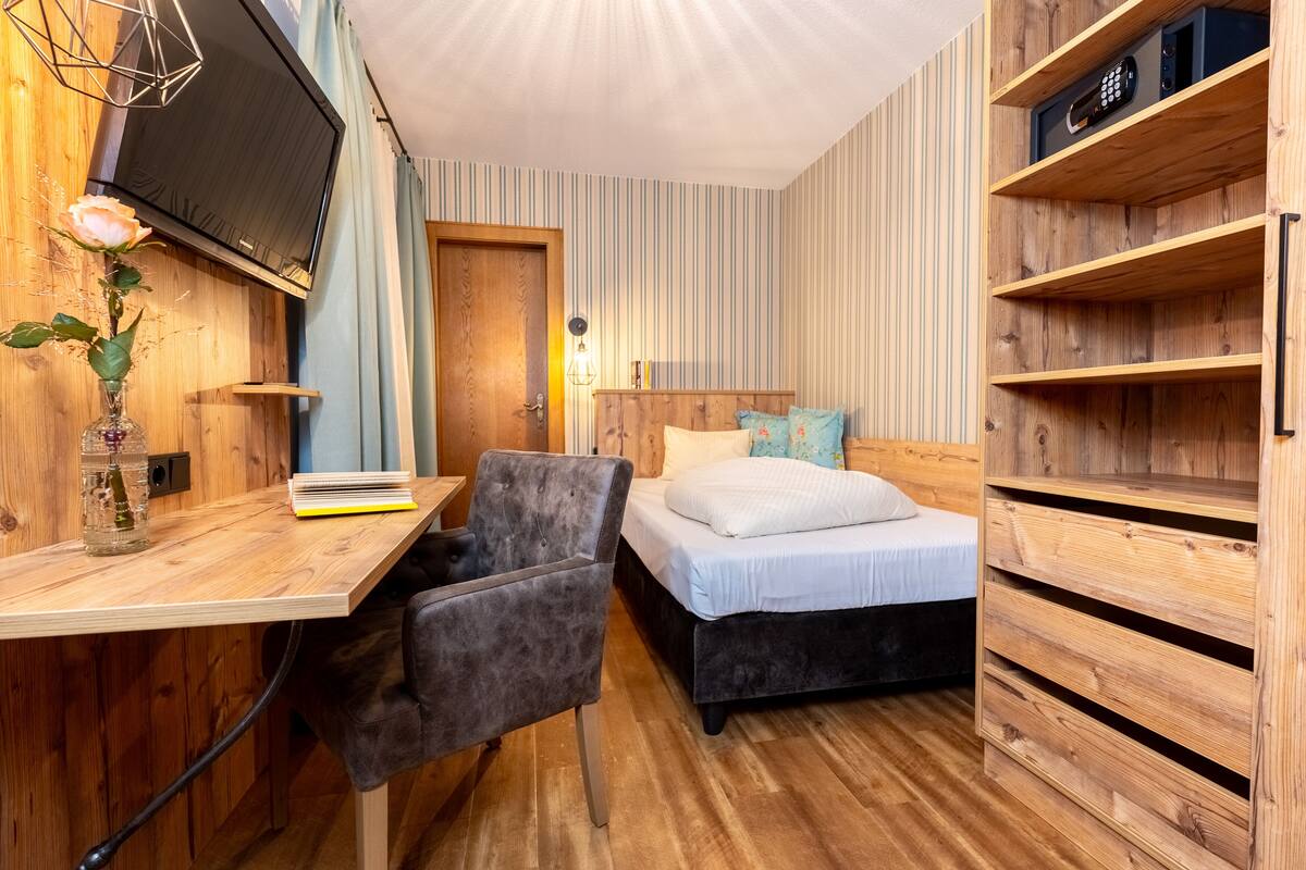 A compact room with a bed, a desk, a chair, and a TV mounted on the wall.