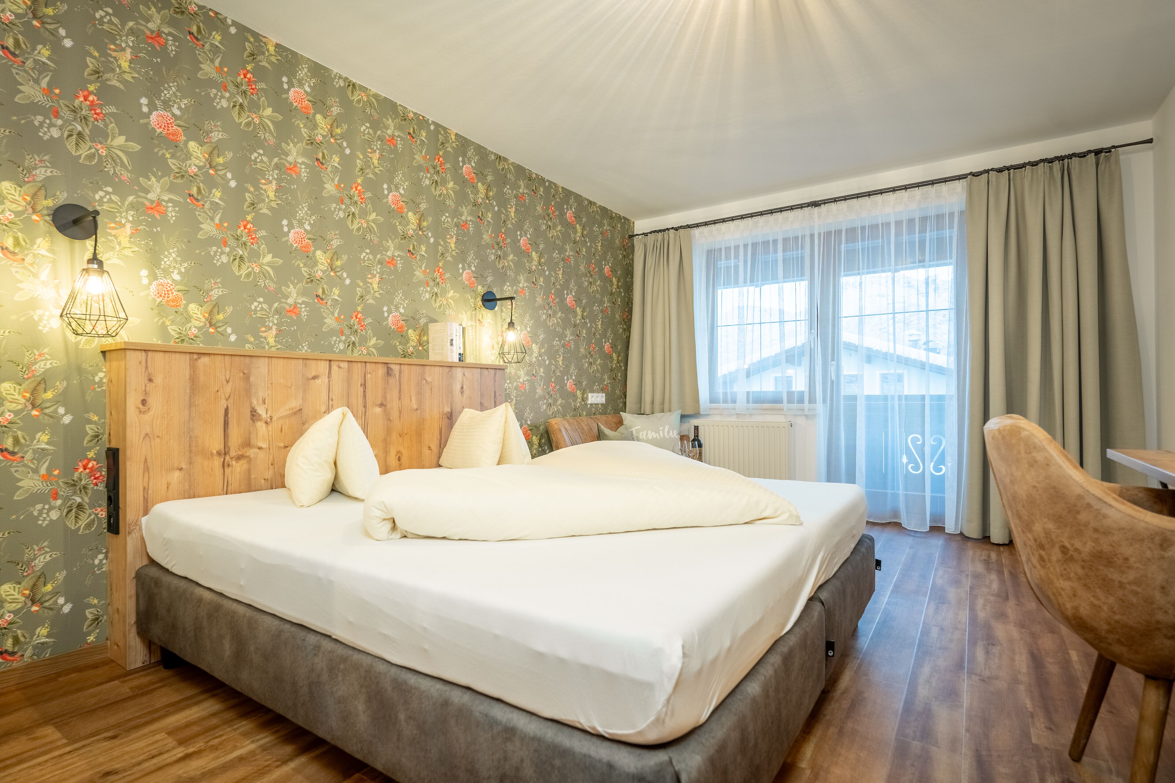 Un dormitorio con cabecera de madera, una cama grande con ropa blanca, papel tapiz floral, una ventana con cortinas translúcidas y una silla de madera.