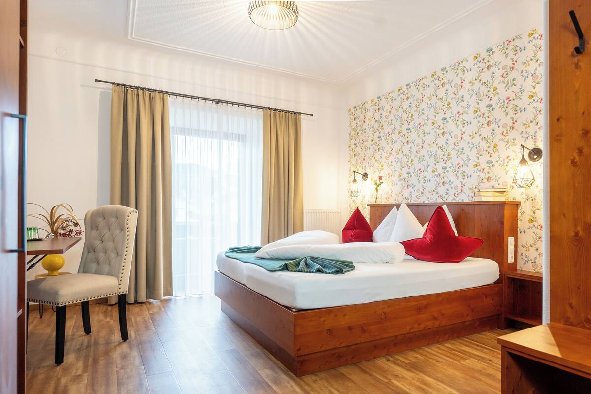 A hotel room with a wooden bed, a chair, a desk, and a window with curtains.