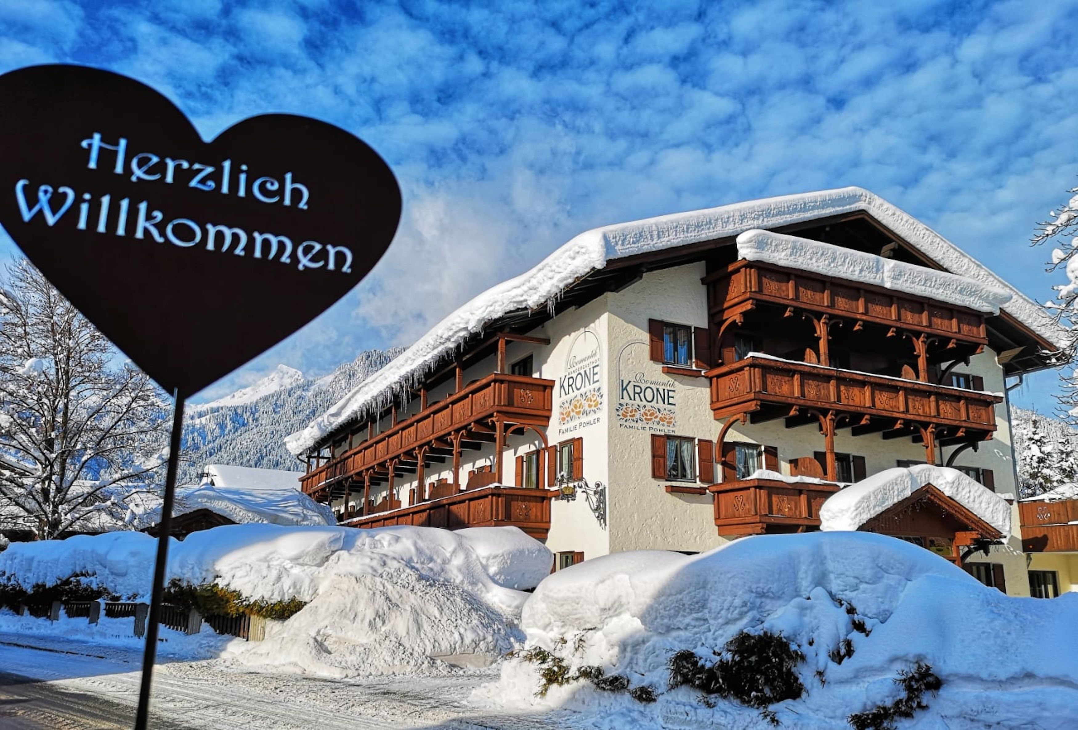A hotel with a heart-shaped sign reading "Herzlich Willkommen" in front of a building with wooden balconies and snow-covered surroundings.
