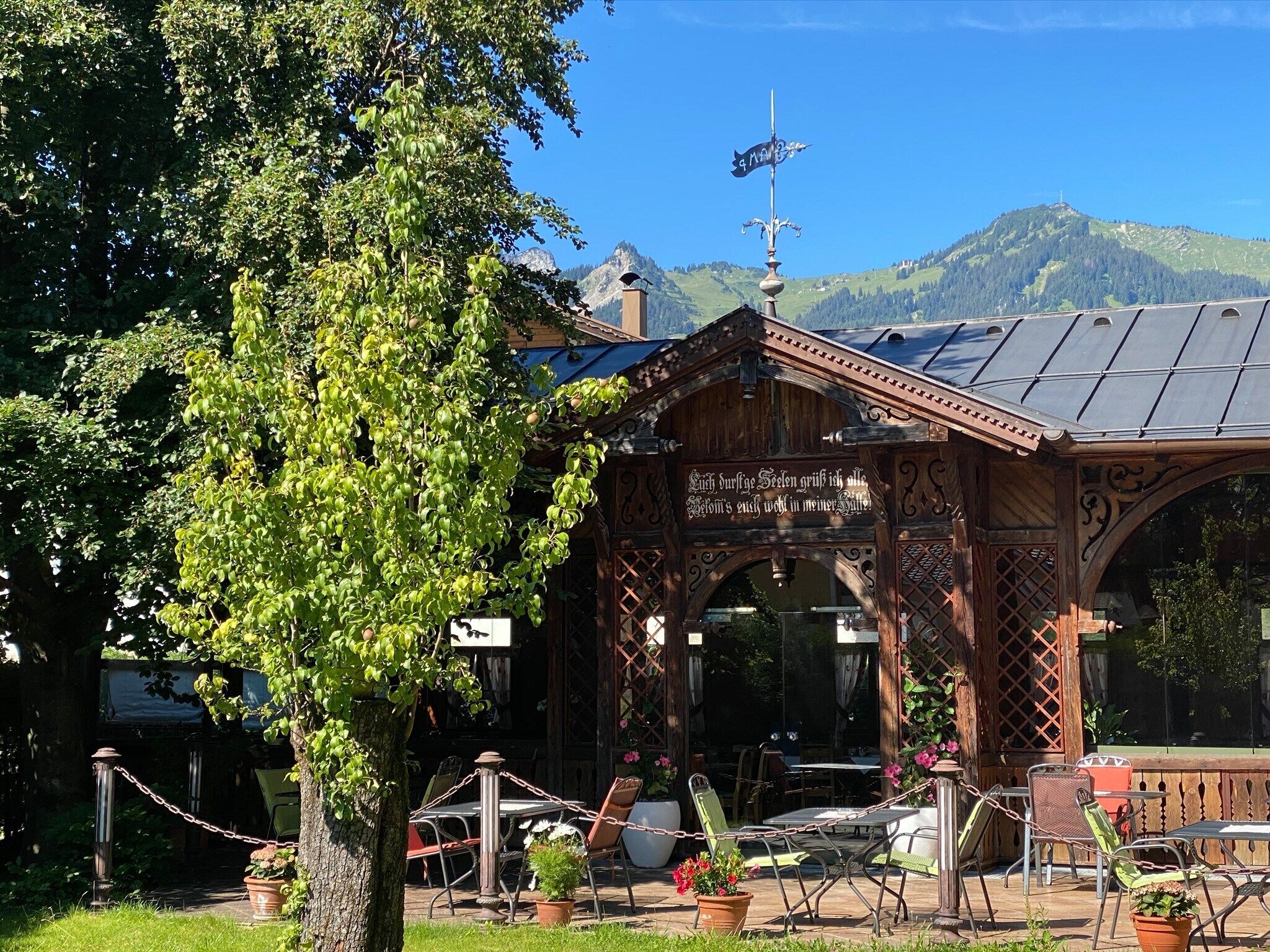 A wooden gazebo with a sign, surrounded by potted plants and outdoor seating.