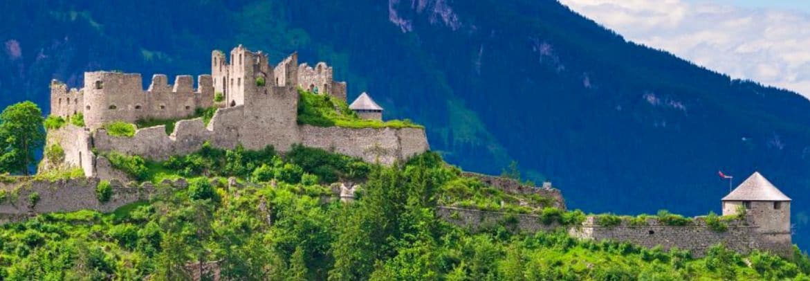 Un château médiéval doté de murs en pierre et de tours, entouré d’une végétation luxuriante et de montagnes.