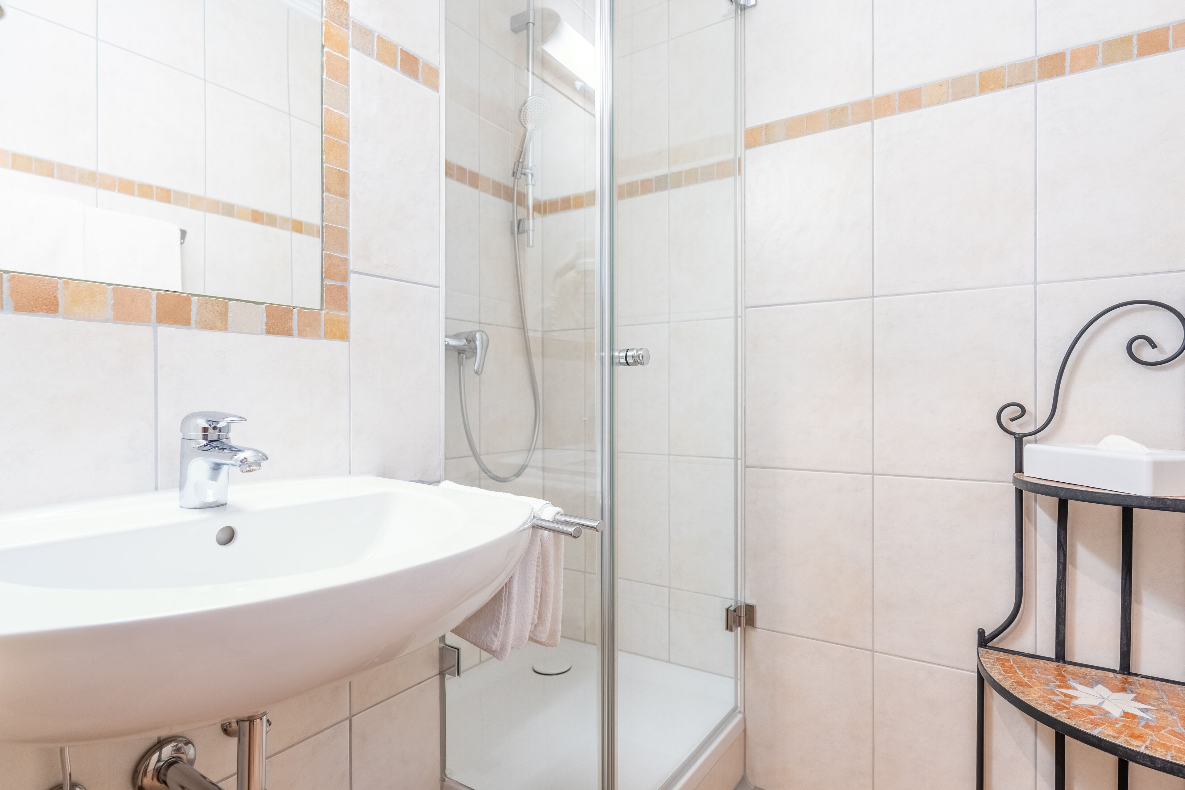 Une salle de bain avec un lavabo blanc, une cabine de douche en verre et un mur carrelé agrémenté d’une bordure décorative.