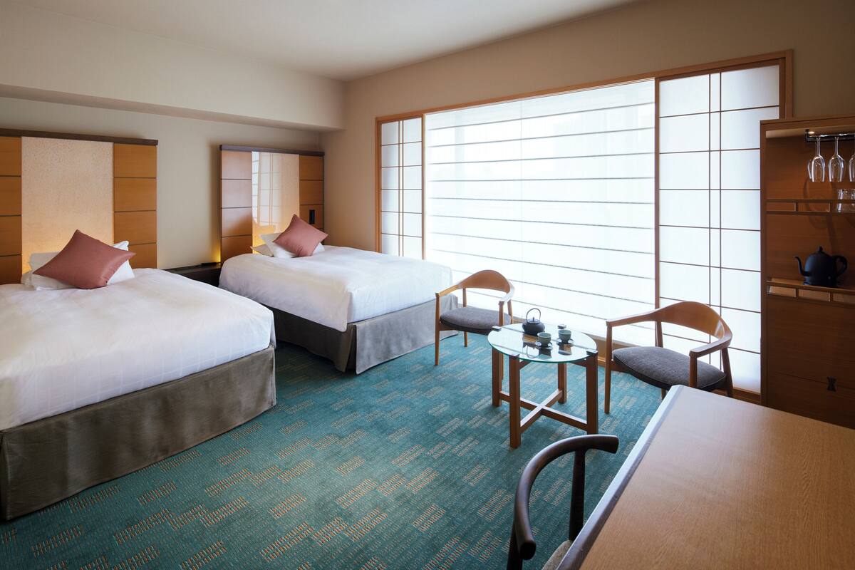 A hotel room with two beds, a small table with chairs, a tea set, and a large window with blinds.