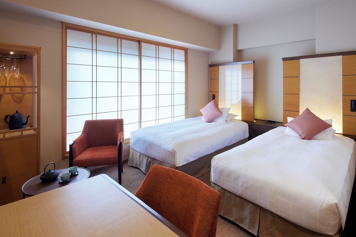 A hotel room with two beds, a sitting area with a table and chairs, a tea set, and a large window with a view.