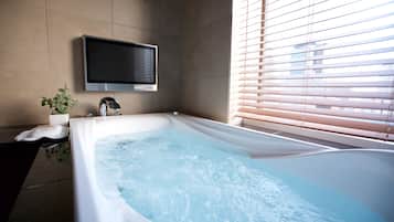 Un baño moderno con una bañera grande, un televisor montado y una ventana con persianas.