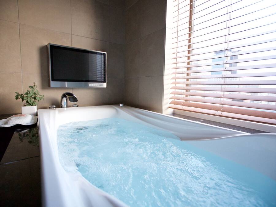 Un baño moderno con una bañera grande, un televisor montado y una ventana con persianas.