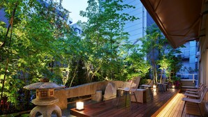 A wooden deck with seating and a stone lantern, surrounded by lush greenery and a water feature.