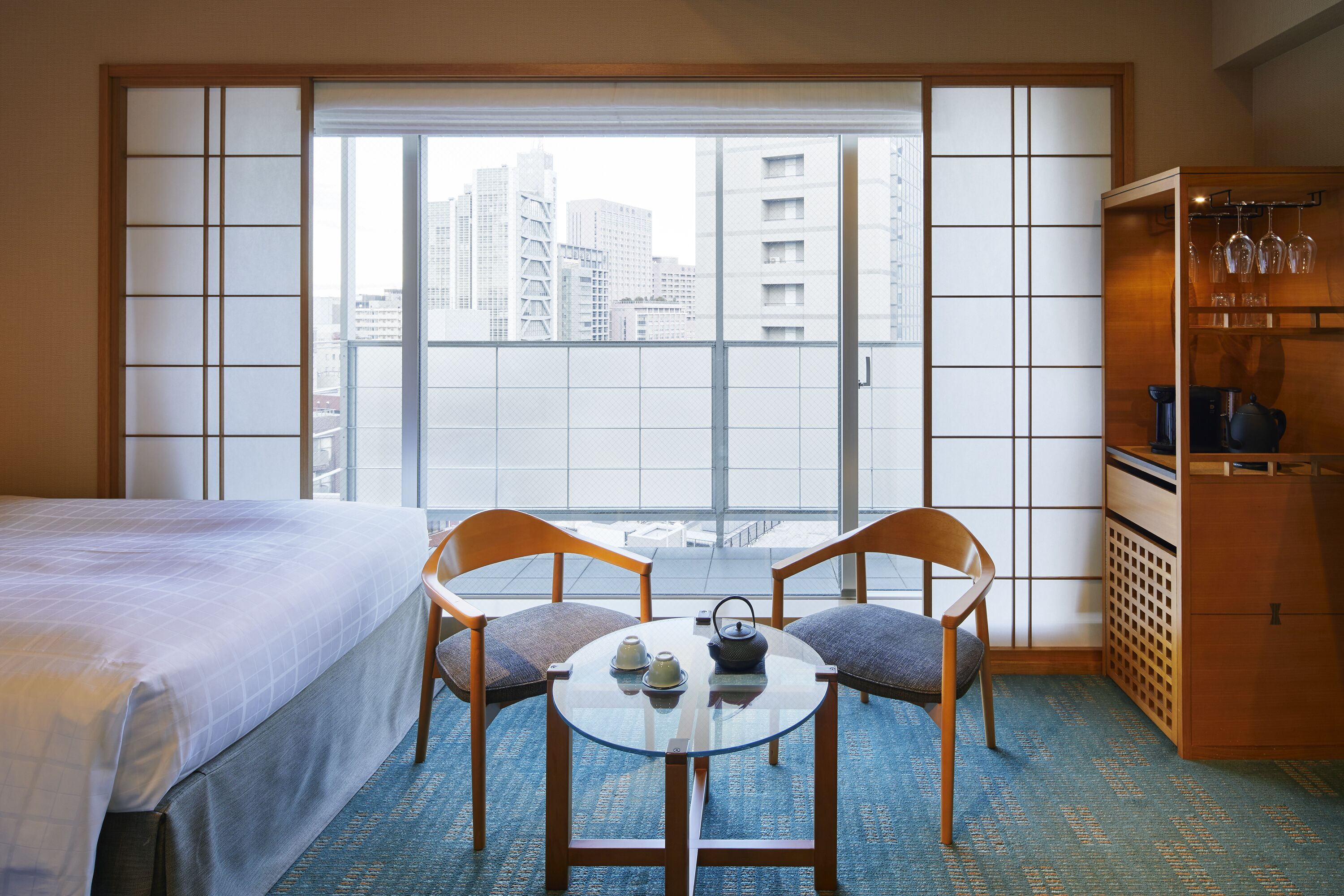 Habitación de hotel con una cama, una mesa redonda de cristal con dos sillas, un juego de té y vistas a edificios altos a través de un celosía japonés.
