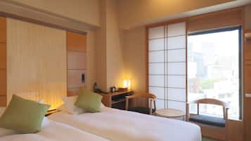 A hotel room with two beds, a desk, a chair, a lamp, and a view of the city through large windows.